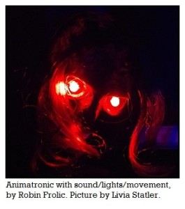 Picture is dark and shows two glowing red eyes shining in a skull face covered with what appears to be dried skin. The eyes are reflecting onto strands of hair that partially cover one eye. The head is tilted. The caption says "Animatronic with sound/lights/movement, by Robin Frolic. Picture taken by Livia Statler."